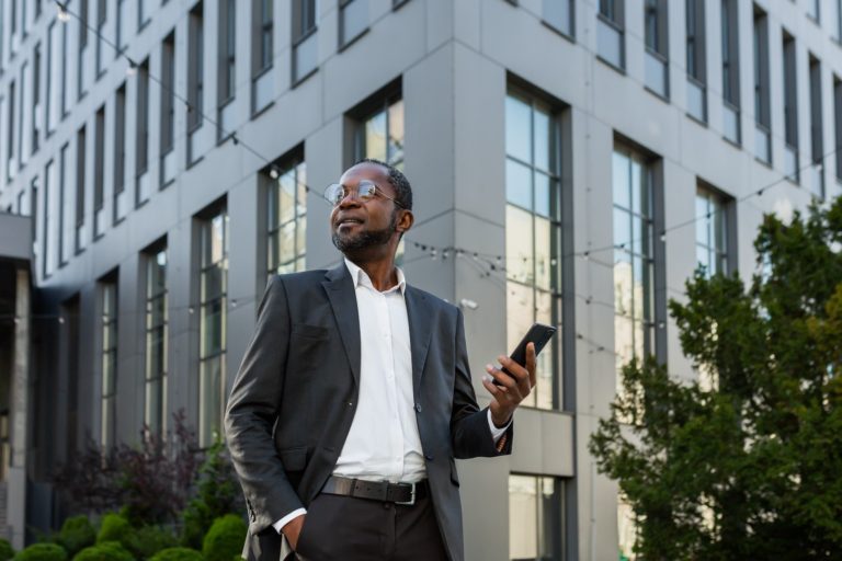 photo from below. portrait of an african american businessman standing in a suit outside an office center and holding a phone. he keeps his hand in his pocket, confidently looks to the side, smiles